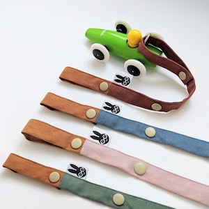 Pu&ograve; includere: Una collezione di clip per ciuccio per bambini colorate in marrone, blu, rosa e verde, ciascuna con chiusura a scatto e un'etichetta con un design a coniglietto. Un'auto giocattolo verde sullo sfondo.