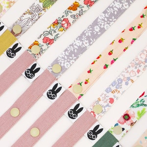 May include: Assortment of colorful fabric straps with a bunny graphic and gold metal hardware. The straps are made of different patterned fabrics, including floral, polka dot, and geometric designs.