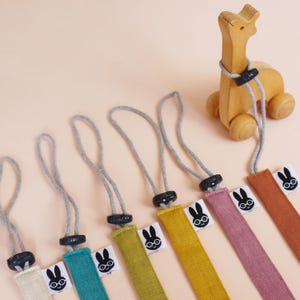 Könnte beinhalten: Eine Auswahl an farbigen Stoff-Lanyards mit einem Hasenlogo, jeweils mit grauem Kordel und schwarzem Verschluss. Ein Holz-Giraffen-Spielzeug zum Ziehen ist ebenfalls im Bild. Die Lanyards sind in Türkis, Gelb, Rosa und Orange gehalten.