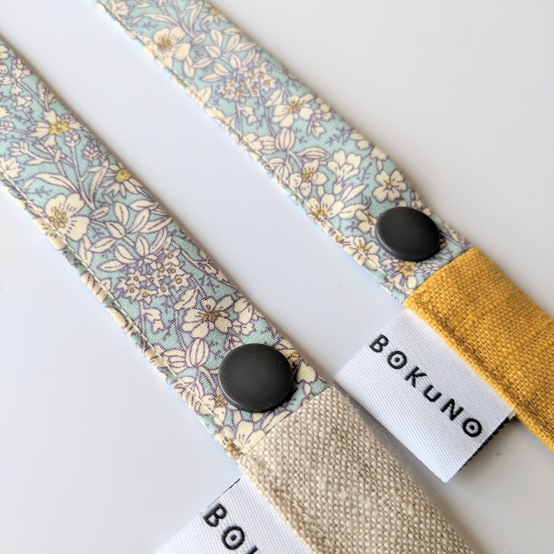 K&ouml;nnte beinhalten: Zwei Stoffb&auml;nder mit Blumenmustern und schwarzen Kn&ouml;pfen. Die B&auml;nder sind an wei&szlig;en Etiketten mit dem in Schwarz gedruckten Text "BOKUNO" befestigt.