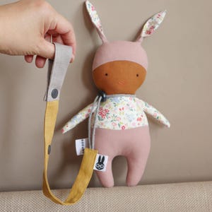 May include: A plush rabbit doll with a brown face, pink head covering, and a floral patterned torso. The doll features long ears with floral lining and is attached to a mustard yellow and gray fabric strap with a snap button and a small tag.