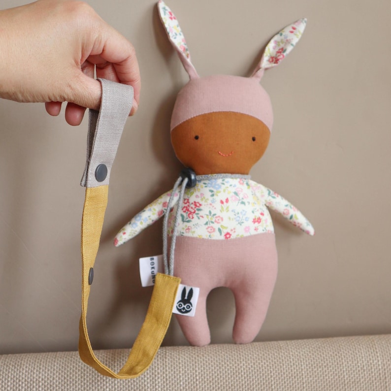 May include: A plush rabbit doll with a brown face, pink head covering, and a floral patterned torso. The doll features long ears with floral lining and is attached to a mustard yellow and gray fabric strap with a snap button and a small tag.