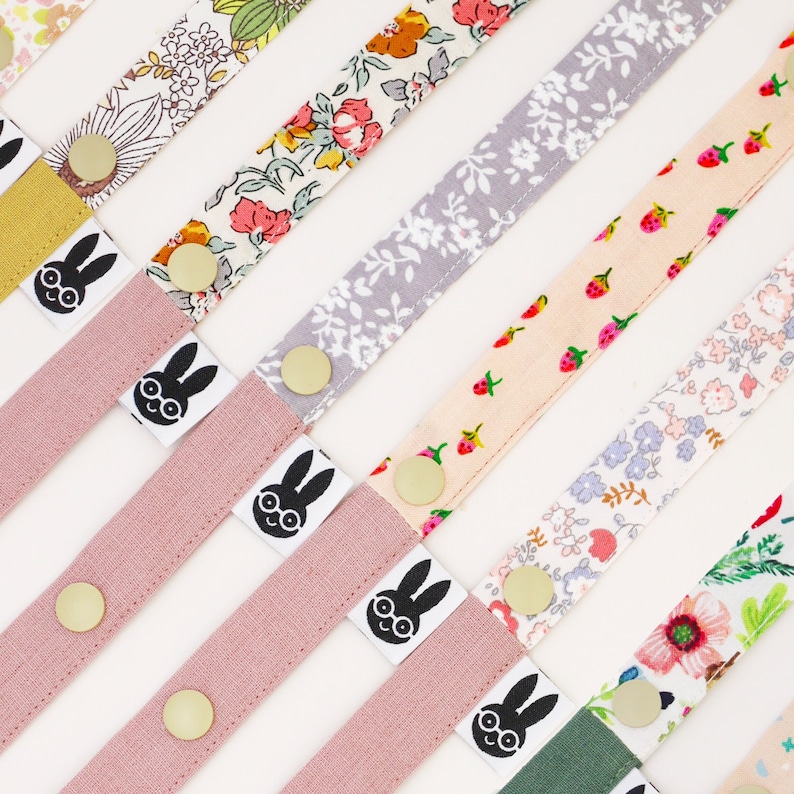 May include: Assortment of colorful fabric straps with a bunny graphic and gold metal hardware. The straps are made of different patterned fabrics, including floral, polka dot, and geometric designs.