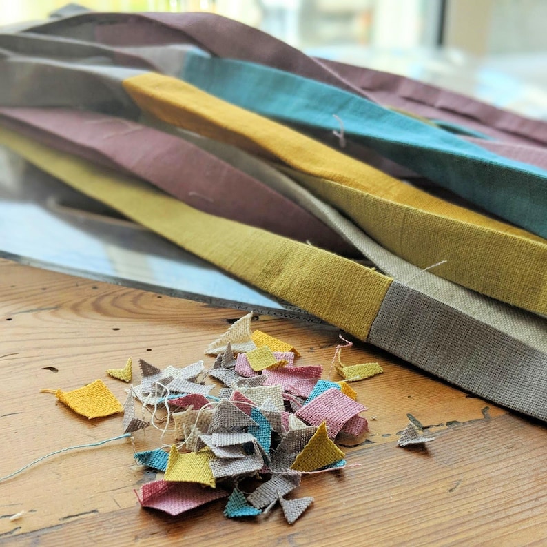 May include: A collection of long, folded fabric strips in shades of mustard yellow, teal, mauve, and gray, accompanied by a scattered pile of small, irregular fabric scraps in matching colors, featuring visible woven textures and loose threads.