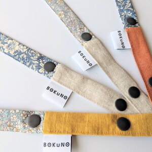 K&ouml;nnte beinhalten: Vier Stoffb&auml;nder mit Blumenmustern und schwarzen Kn&ouml;pfen. Jedes Band hat ein wei&szlig;es Etikett mit dem aufgedruckten Text "BOKUNO".