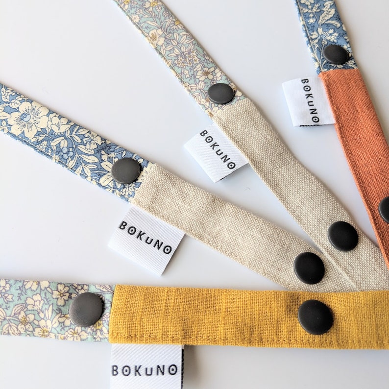 K&ouml;nnte beinhalten: Vier Stoffb&auml;nder mit Blumenmustern und schwarzen Kn&ouml;pfen. Jedes Band hat ein wei&szlig;es Etikett mit dem aufgedruckten Text "BOKUNO".