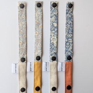 K&ouml;nnte beinhalten: Vier Stoffb&auml;nder mit Blumenmustern und schwarzen verstellbaren Schnallen. Jedes Band hat eine andere farbige Stoffr&uuml;ckseite: Naturleinen, Senfgelb, Naturleinen und gebranntes Orange. Die B&auml;nder sind mit "BOKUNO" beschriftet.