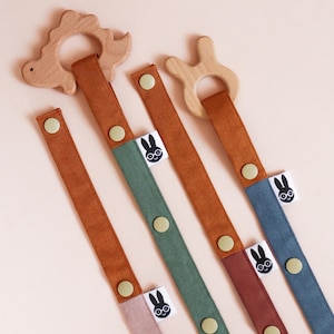 May include: A collection of baby pacifier clips in various colors. Each clip features a wooden teething ring in the shape of a dinosaur or a bunny. The straps are in shades of brown, green, blue, and pink, with a button closure and a label with a bunny logo.