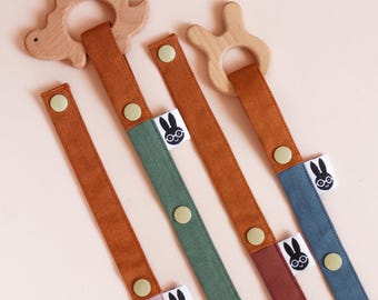 May include: A collection of baby pacifier clips in various colors. Each clip features a wooden teething ring in the shape of a dinosaur or a bunny. The straps are in shades of brown, green, blue, and pink, with a button closure and a label with a bunny logo.