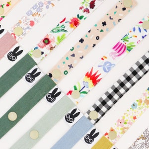 Könnte beinhalten: Sortiment an bunten Stoffbändern mit verschiedenen Mustern, darunter florale, geometrische und karierte Designs. Jedes Band zeigt eine schwarz-weiße Grafik eines Kaninchens mit Brille. Die Bänder sind etwa 30 cm lang.