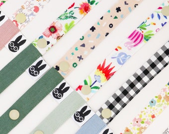 May include: Assortment of colorful fabric straps with different patterns, including floral, geometric, and checkered designs. Each strap features a black and white graphic of a bunny with glasses. The straps are approximately 12 inches long.