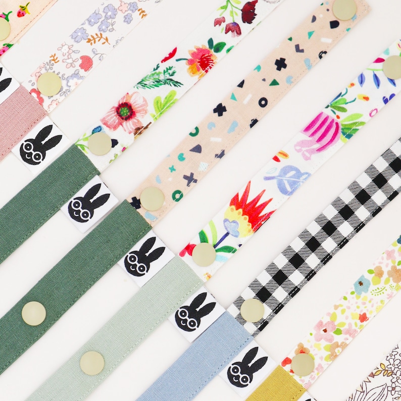 May include: Assortment of colorful fabric straps with different patterns, including floral, geometric, and checkered designs. Each strap features a black and white graphic of a bunny with glasses. The straps are approximately 12 inches long.