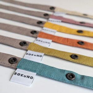 May include: A collection of fabric straps in various colors including teal, mustard yellow, orange, pink, and beige. Each strap features a dark gray snap button and a white fabric tag with the black text BOKUNO printed on it.