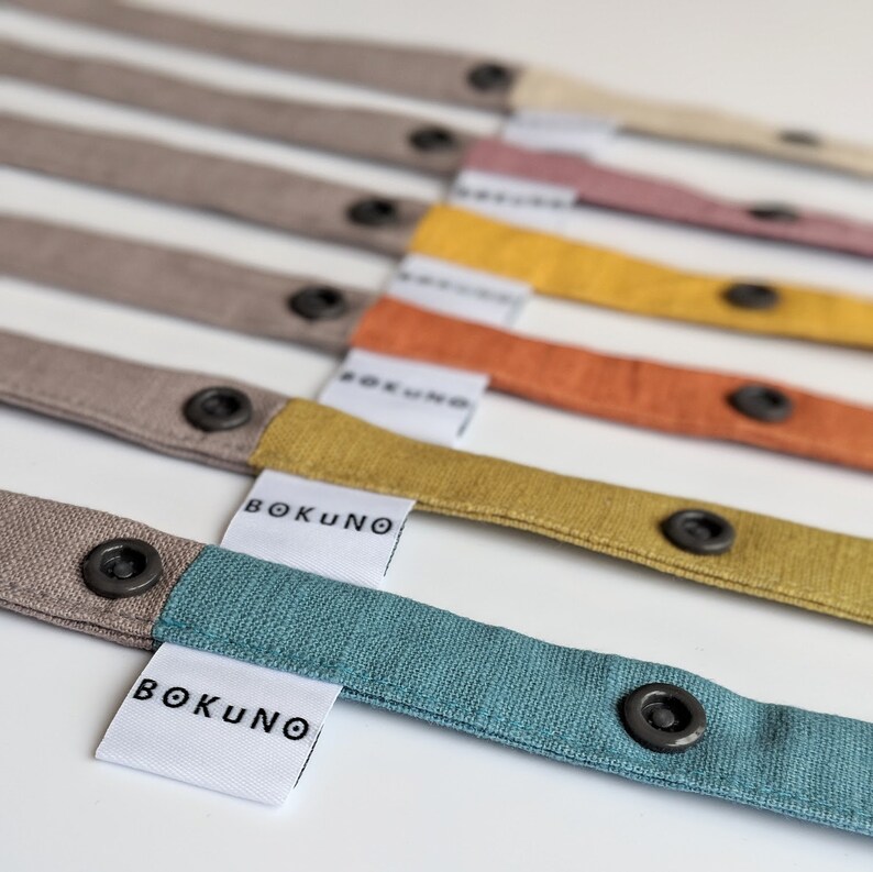 May include: A collection of fabric straps in various colors including teal, mustard yellow, orange, pink, and beige. Each strap features a dark gray snap button and a white fabric tag with the black text BOKUNO printed on it.