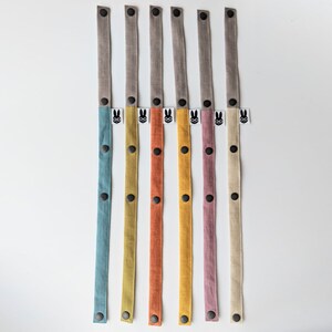 May include: Six long fabric straps with snap buttons, each featuring a grey top section and a colored bottom section in blue, olive, orange, yellow, mauve, or beige. Each strap includes a small white tag with a black rabbit icon wearing glasses.