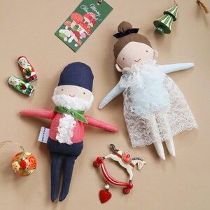 May include: A selection of Christmas decorations, including fabric dolls and festive ornaments. One doll is dressed in a red coat and blue trousers, the other in a blue dress with lace. Other decorations include a rocking horse, a Christmas tree, and a tag that says "Merry Christmas!"
