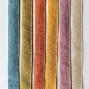 May include: Six vertical fabric strips in a row, featuring a textured weave. The colors from left to right are muted blue, olive green, burnt orange, golden yellow, dusty rose, and natural beige. Each strip has stitched edges along both sides.