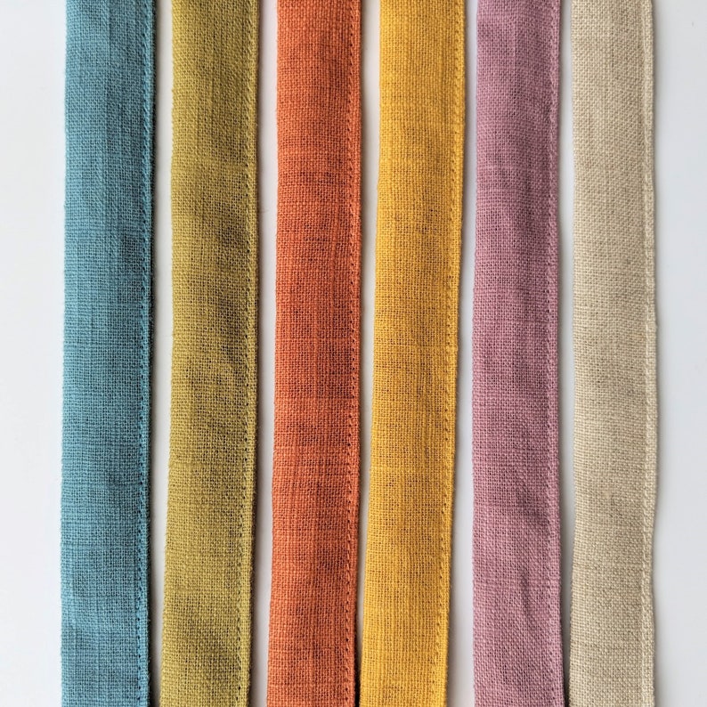 May include: Six vertical fabric strips in a row, featuring a textured weave. The colors from left to right are muted blue, olive green, burnt orange, golden yellow, dusty rose, and natural beige. Each strip has stitched edges along both sides.