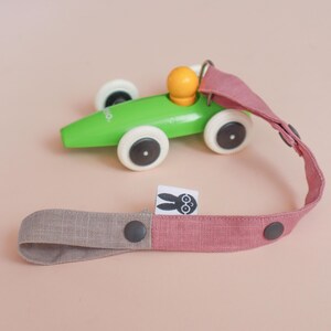 May include: A bright green wooden toy race car with white wheels and a yellow driver figure, attached to a long, two-toned dusty pink and beige fabric strap. The strap features dark gray snap buttons and a small white tag with a black rabbit icon.