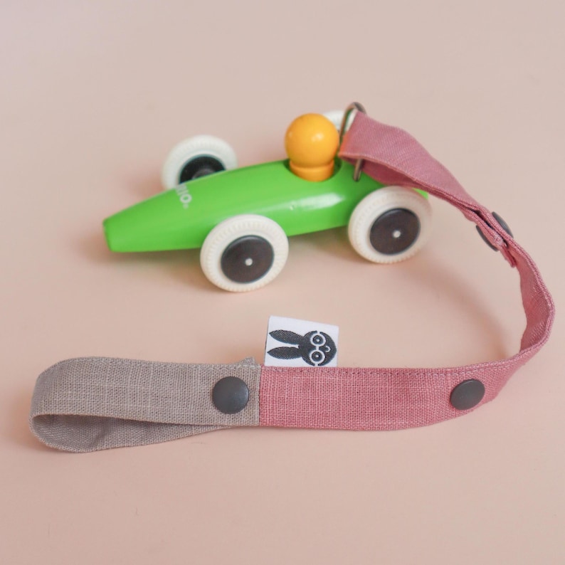 May include: A bright green wooden toy race car with white wheels and a yellow driver figure, attached to a long, two-toned dusty pink and beige fabric strap. The strap features dark gray snap buttons and a small white tag with a black rabbit icon.