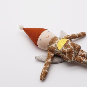 May include: A fabric doll with a rust-coloured pointed hat topped with a white pom-pom, wearing a tan outfit with a brown oak leaf pattern. The doll features a gold coin tucked into a front pocket and has light grey fabric wings on its back.