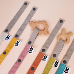May include: A collection of fabric pacifier clips featuring long, multi-colored linen straps in yellow, pink, orange, and teal, each with dark gray snap buttons and a small white tag displaying a black rabbit icon, attached to wooden teethers.