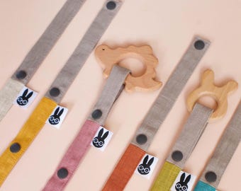 May include: A collection of fabric pacifier clips featuring long, multi-colored linen straps in yellow, pink, orange, and teal, each with dark gray snap buttons and a small white tag displaying a black rabbit icon, attached to wooden teethers.