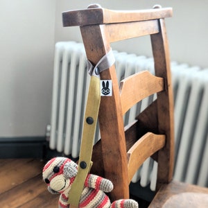 May include: A mustard yellow fabric strap with black snap buttons and a small white tag featuring a black rabbit icon with glasses. The strap is looped over a wooden chair and holds a crocheted monkey toy with red, white, and gray stripes.