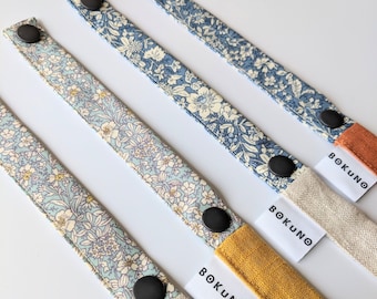 May include: Four fabric straps with floral patterns and black buttons. Each strap has a white label with the word "BOKUNO" printed on it. The straps are attached to different colored fabric pieces: orange, yellow, and beige.