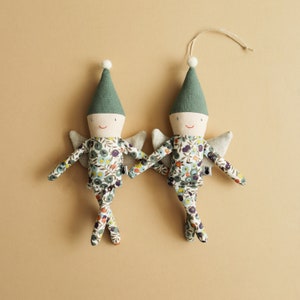 May include: Two fabric elf dolls with green pointed hats and white bobbles. The dolls have smiling faces, small wings, and floral patterned outfits. They are holding hands and have a string for hanging.