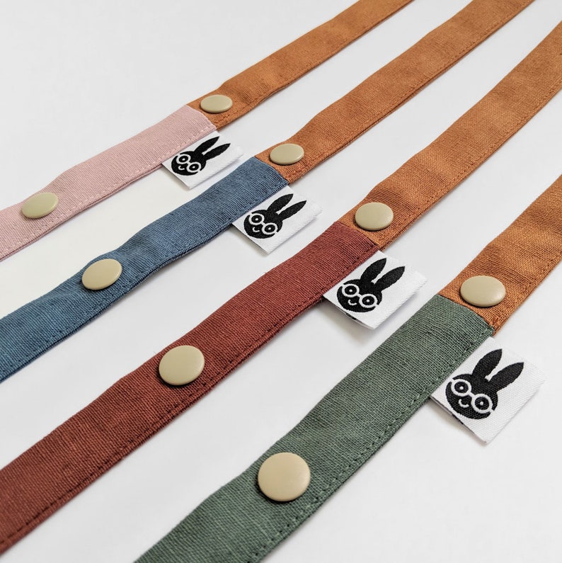 Pu&ograve; includere: Primo piano di cinghie in tessuto di vari colori: rosa, blu, ruggine, marrone e verde. Ogni cinghia ha un bottone e una piccola etichetta bianca con un volto di coniglio nero. Le cinghie sono disposte diagonalmente su sfondo bianco.