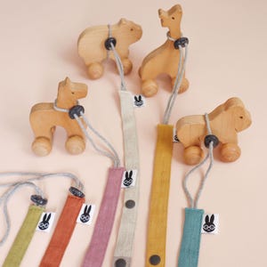 May include: A collection of wooden animal figures on wheels, each attached to a long fabric strap in various colors including mustard, teal, pink, and green. Each strap features a small white tag with a black rabbit icon and a dark button.