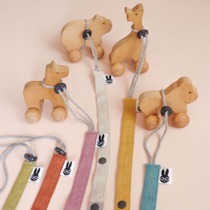 May include: A collection of wooden animal figures on wheels, each attached to a long fabric strap in various colors including mustard, teal, pink, and green. Each strap features a small white tag with a black rabbit icon and a dark button.