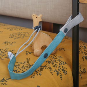 May include: A wooden giraffe toy on wheels stands next to a teal and gray fabric strap with dark gray snap buttons. The strap features a small white tag with the text BOKUNO printed in black. The toy has a smooth, light brown finish.