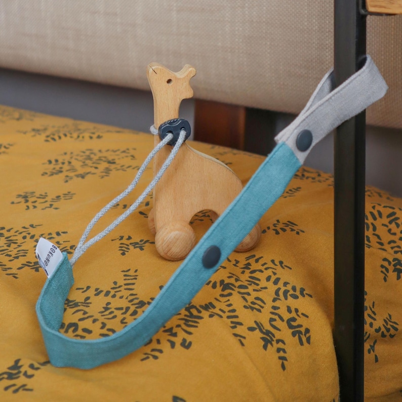 May include: A wooden giraffe toy on wheels stands next to a teal and gray fabric strap with dark gray snap buttons. The strap features a small white tag with the text BOKUNO printed in black. The toy has a smooth, light brown finish.