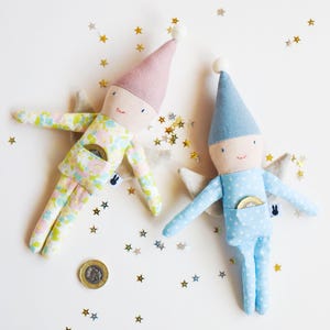 May include: Two handmade fabric dolls with star-shaped wings and pointed hats. One doll is pink and floral, the other is blue with white polka dots. Both have a small pocket and a coin. Gold star confetti is scattered around.