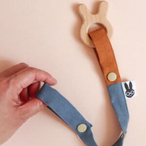 Pu&ograve; includere: Un massaggiagengive per bambini in legno con un anello a forma di coniglio e un cinturino in tessuto blu e arancione. Il cinturino ha una chiusura a bottone e una piccola etichetta con un logo di coniglio. Il massaggiagengive &egrave; tenuto in mano su sfondo rosa chiaro.
