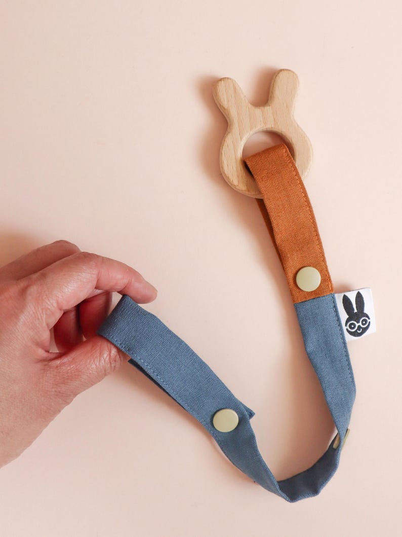 Pu&ograve; includere: Un massaggiagengive per bambini in legno con un anello a forma di coniglio e un cinturino in tessuto blu e arancione. Il cinturino ha una chiusura a bottone e una piccola etichetta con un logo di coniglio. Il massaggiagengive &egrave; tenuto in mano su sfondo rosa chiaro.
