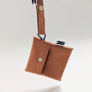 Pu&ograve; includere: Una piccola pochette color ruggine con chiusura a patta assicurata da un bottone a pressione dorato. Un cinturino abbinato &egrave; attaccato alla pochette con un cordino nero. La pochette ha una piccola etichetta bianca con una grafica di coniglietto nero.