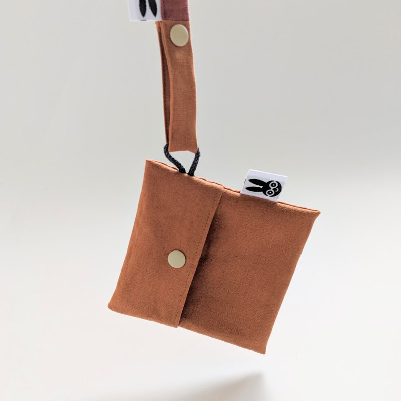 Pu&ograve; includere: Una piccola pochette color ruggine con chiusura a patta assicurata da un bottone a pressione dorato. Un cinturino abbinato &egrave; attaccato alla pochette con un cordino nero. La pochette ha una piccola etichetta bianca con una grafica di coniglietto nero.