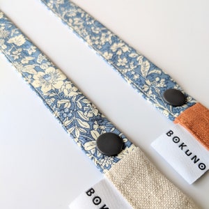 K&ouml;nnte beinhalten: Zwei blaue und wei&szlig;e Blumenstoffb&auml;nder mit schwarzen Kn&ouml;pfen und einem wei&szlig;en Etikett mit der Aufschrift "BOKUNO".