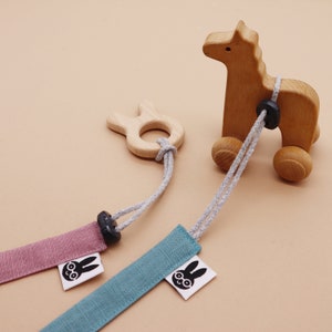 May include: A wooden horse toy on wheels connected by grey cords to two fabric straps in dusty pink and teal. Each strap features a white tag with a black graphic of a rabbit wearing glasses. A small wooden rabbit-shaped ring is attached to one cord.