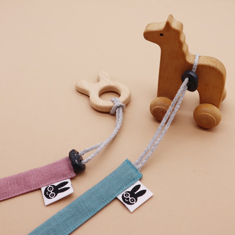 May include: A wooden horse toy on wheels connected by grey cords to two fabric straps in dusty pink and teal. Each strap features a white tag with a black graphic of a rabbit wearing glasses. A small wooden rabbit-shaped ring is attached to one cord.