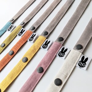 May include: A collection of fabric straps in various colors including teal, mustard yellow, orange, pink, and off-white. Each strap features a dark gray snap button and a white fabric tag displaying a black graphic of a rabbit wearing glasses.
