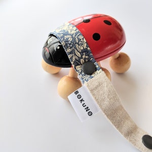 K&ouml;nnte beinhalten: Ein rotes und schwarzes Marienk&auml;fer-Spielzeug mit Holzr&auml;dern und einem blauen und wei&szlig;en floralen Stoffband. Das Band hat ein wei&szlig;es Etikett mit dem Text "BOKUNO" darauf.