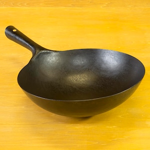 May include: A black wok with a long handle. The wok is sitting on a wooden surface.