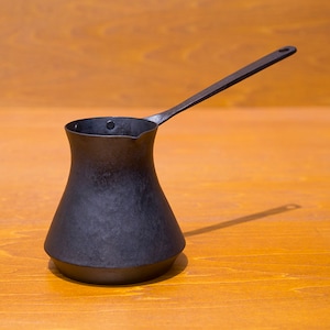 Puede incluir: Una cafetera de metal negro con un mango largo. La cafetera está sobre una superficie de madera.