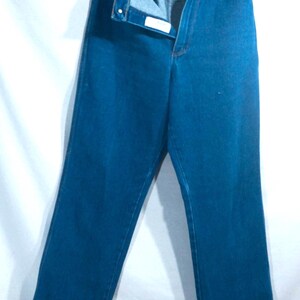 May include: A pair of blue denim jeans hanging from a hanger. The jeans are unbuttoned and unzipped, revealing the interior. The denim is a solid, medium blue color. The jeans have pockets and a button closure.