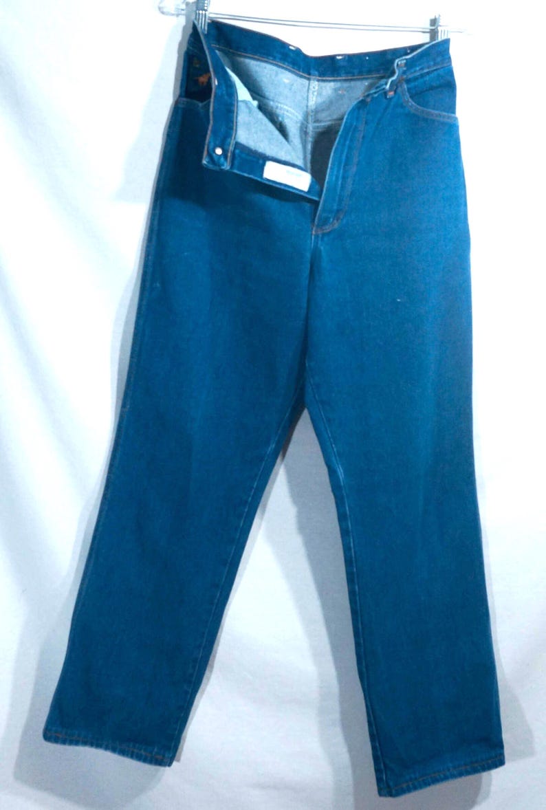 May include: A pair of blue denim jeans hanging from a hanger. The jeans are unbuttoned and unzipped, revealing the interior. The denim is a solid, medium blue color. The jeans have pockets and a button closure.