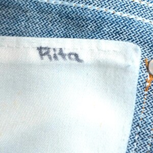 May include: Close-up of denim jeans with a white fabric label. The word "Rita" is printed in black on the label. A silver button is visible on the jeans.